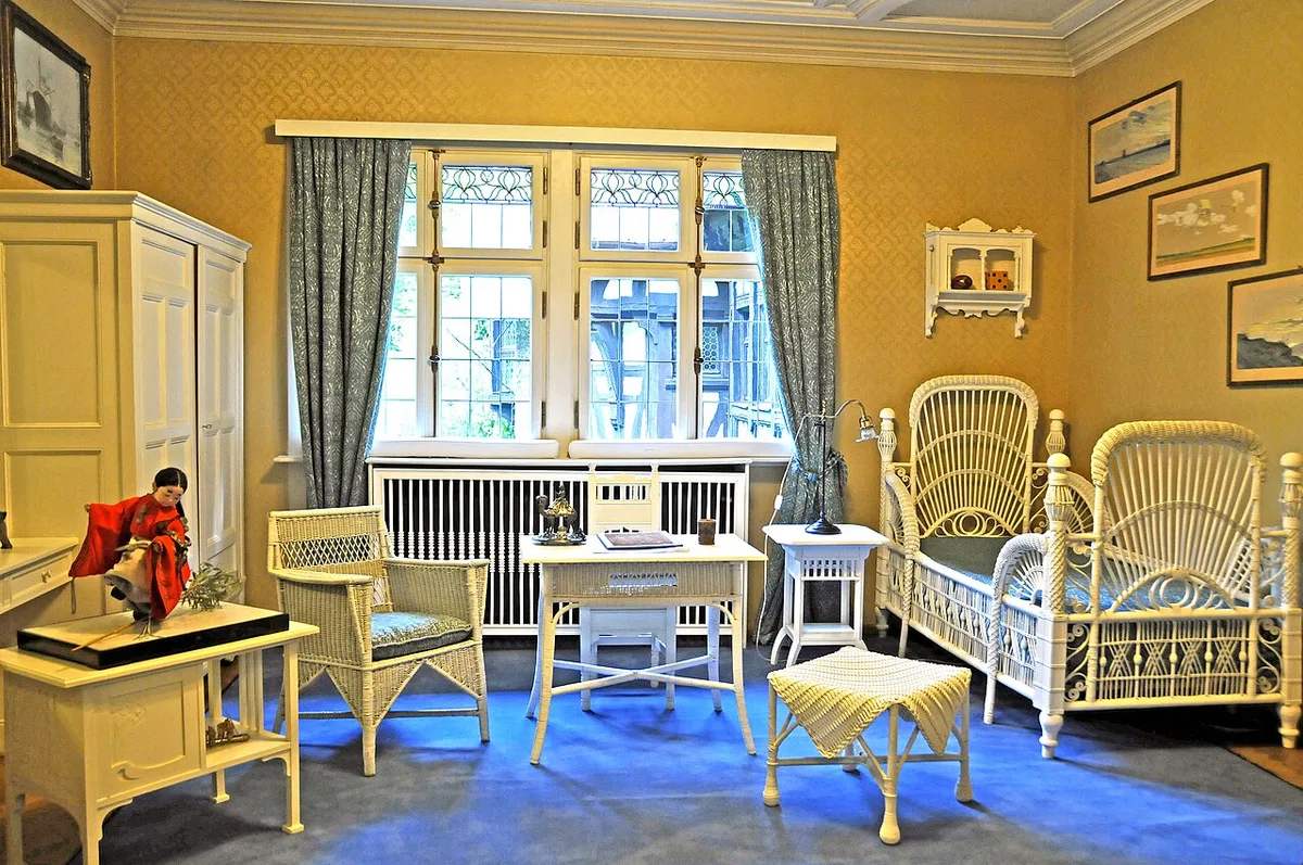 Kinderkamer met gele wanden en rieten meubels, met een wandplankje en ingelijste prenten boven het bed.