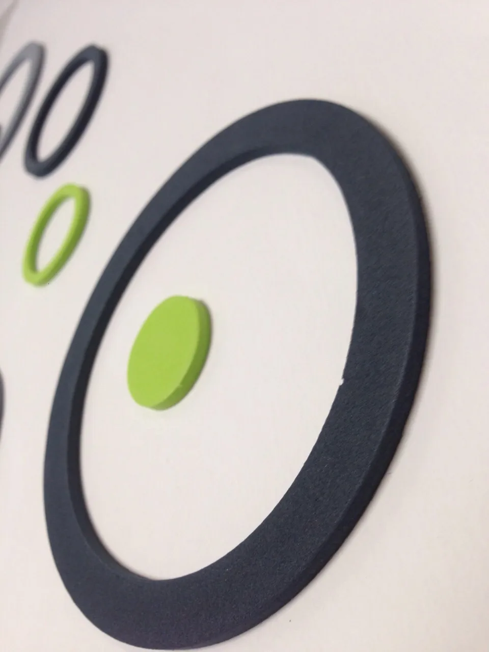 Detail van een driedimensionale muursticker van rubber: een donkere ring met groene stippen op een witte wand.