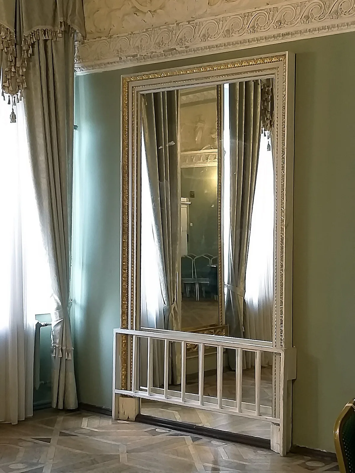 Grote wandspiegel in een witte lijst die een zaal met lichte gordijnen en hoge ramen weerspiegelt.